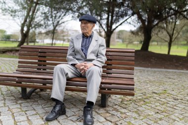 Takım elbiseli ve düz şapkalı yaşlı bir beyefendi bir bankta oturuyor, düşüncelere dalmış.