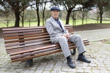 Takım elbiseli ve şapkalı yaşlı bir adam parktaki bankta oturuyor, düşünceli ve düşünceli görünüyor.