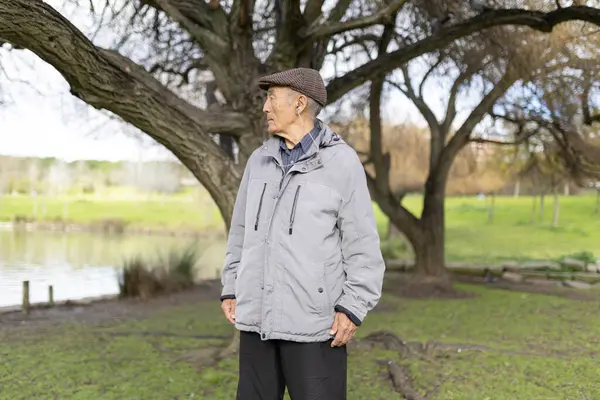 Şapka ve ceket giyen yaşlı bir adam gölün kenarındaki parkta huzurlu bir yürüyüşün tadını çıkarıyor.