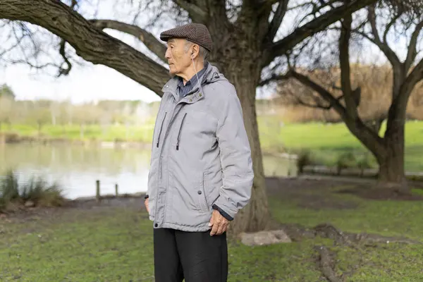 Gri ceketli ve şapkalı yaşlı bir adam huzur dolu bir parkta göl kenarında yürürken müzik dinliyor.