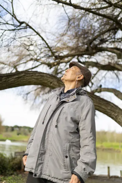 Yaşlı bir adam gölün yakınındaki bir parkta ağaçlara bakarken kulaklıkla müzik dinliyor.