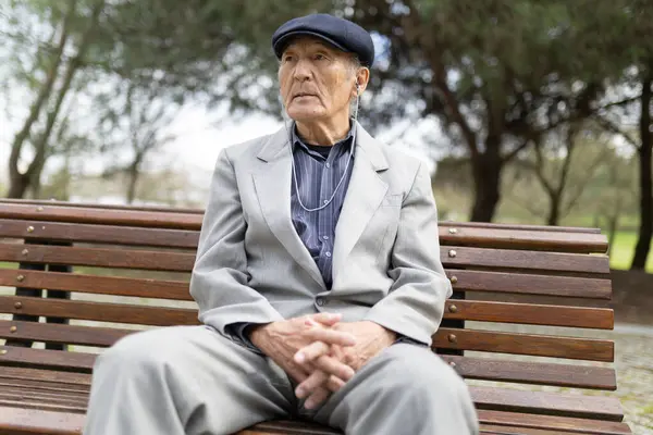 Son sınıf öğrencisi şık bir takım elbise ve şapka giyip parkta bankta otururken kulaklıkla müzik dinliyor.