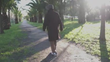 Kahverengi ceketli bir adam güneşli bir günde parktaki palmiye ağaçları arasındaki yolda yürüyor.