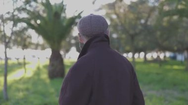 Kahverengi ceketli ve düz şapkalı son sınıf öğrencisi yeşil bir parkta huzurlu bir yürüyüşün tadını çıkarıyor.