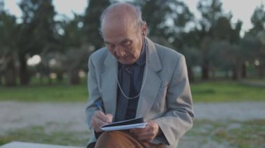 Son sınıf öğrencisi bankta oturuyor, parkta not defterine yazıyor.