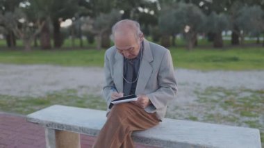 Not defterine yazan yaşlı bir adam parkta bankta oturuyor.