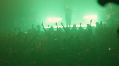 Yeşil ışıklar ve sis bir konser salonunu dolduruyor. Şarkıcı sahnede şarkı söylerken kalabalık tezahürat yapıyor ve ellerini kaldırıyor.