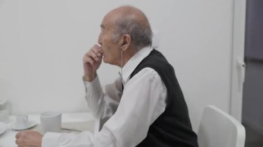 İşitme cihazlı kıdemli adam huzur dolu bir kahvaltıyı seviyor, küçük lokmalar yiyip kahvenin tadını çıkarıyor.