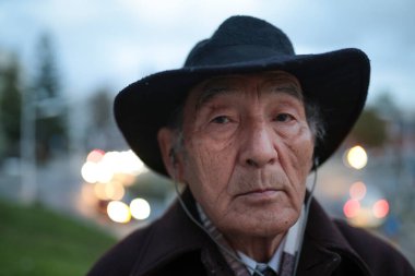 Şapka ve kulaklık takan yaşlı bir Asyalı 'nın portresi, alacakaranlıkta şehir ortamında duruyor.