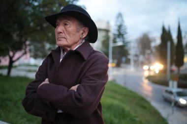 Akşamleyin yolun kenarındaki parkta duran kollarını çapraz bağlayan şapkalı dalgın bir son sınıf öğrencisi.