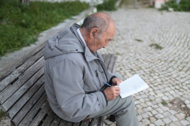 Kulaklık kullanan kıdemli bir adam, parkta bir bankta otururken deftere not alıyor.