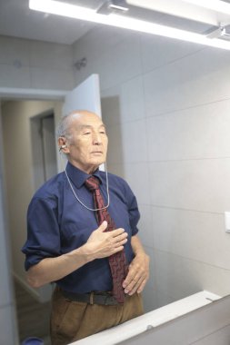 Yaşlı işadamı banyoda hazırlanıyor, işitme cihazını takarken kravatını düzeltiyor.