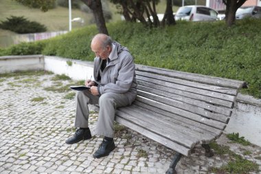Yaşlı profesör bankta otururken not defterine notlar yazıyor, temiz havanın ve sessiz ortamın tadını çıkarıyor.