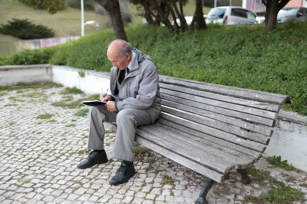 Yaşlı profesör bankta otururken not defterine notlar yazıyor, temiz havanın ve sessiz ortamın tadını çıkarıyor.