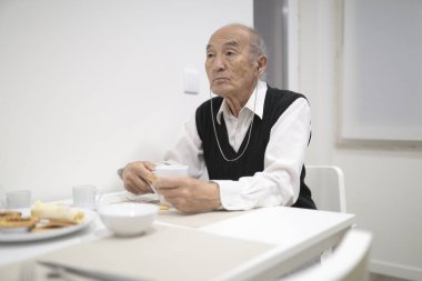 İşitme cihazı takan son sınıf öğrencisi, beyaz bir yemek odasında kahvaltı yapıyor.