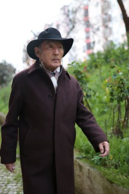 Üst düzey bir adam parkta rahatça yürüyor, şapka, ceket ve kulaklık giyiyor, doğanın huzurunu kucaklıyor.