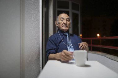 Kulaklıklı yaşlı bir adam ve bir fincan kahve geceleri balkonda oturuyor.