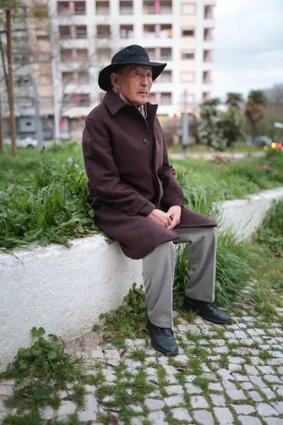 Sessiz bir şehir parkında, alçak bir duvarda oturan şapka ve palto giyen yaşlı bir adam, temiz havanın ve şehrin tadını çıkarıyor.