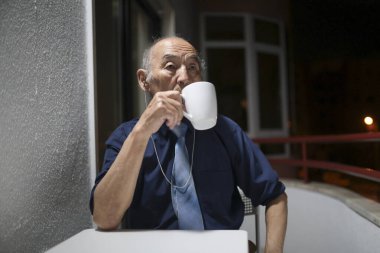 Yaşlı işadamı geceleri balkonunda kahve içip kulaklıkla müzik dinliyor.
