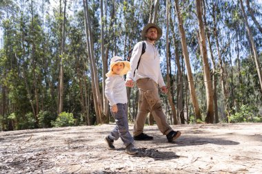 Baba ve oğul güneşli bir ormanda el ele tutuşarak ve şapka takarak sakin bir yürüyüş yapıyorlar.