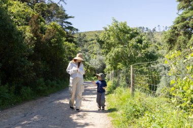 Anne ve oğlu toprak bir yolda yürüyor, güneşli bir günün tadını çıkarıyorlar.