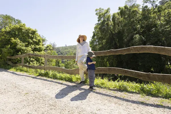 Anne ve oğlu yürüyüş sırasında mola veriyor, vadiye bakan ahşap çitlere yaslanıyorlar.