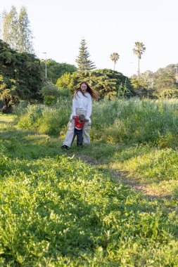 Anne oğlunu güneşli bir bahar gününde parkta çayırda yürürken taşıyor.