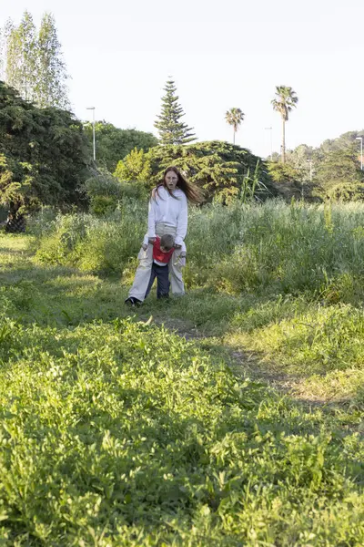 Anne oğlunu güneşli bir bahar gününde parkta çayırda yürürken taşıyor.