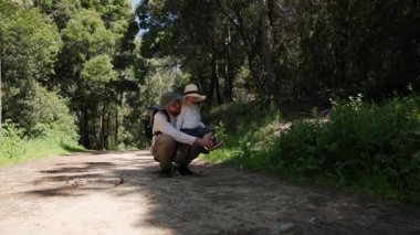 Baba, güneşli bir yolda yürürken küçük çocuğuna ayakkabılarını bağlamasında yardım ediyor.