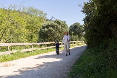 Anne ve oğlu güneşli bir bahar gününde, yemyeşil ve ahşap çitlerle çevrili, sakin bir yürüyüşün tadını çıkarıyorlar.