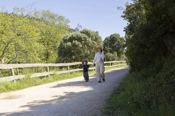 Anne ve oğlu güneşli bir bahar gününde, yemyeşil ve ahşap çitlerle çevrili, sakin bir yürüyüşün tadını çıkarıyorlar.