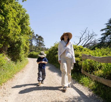 Şapka takan anne ve oğul güneşli bir yaz gününde doğa koruma alanında çakıllı bir yolda yürüyorlar.