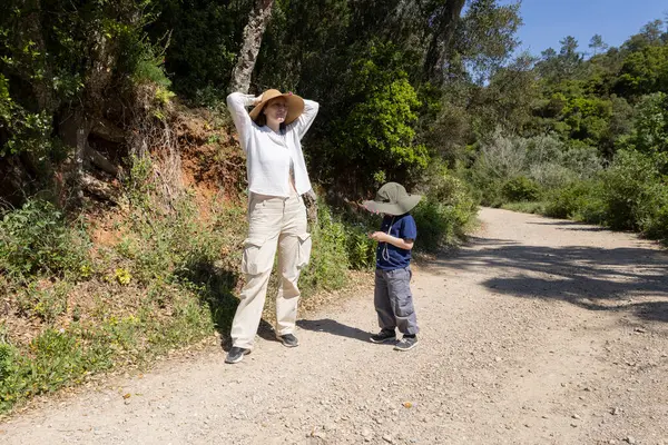 Güneşli bir yaz gününde, şapka takan anne ve oğul doğa yürüyüşü yapıyorlar.