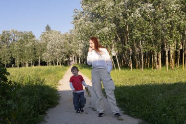 Anne ve oğlu güneşli bir yaz gününde birlikte yürüyüş yapıyor, yemyeşil ağaçların arasında yol boyunca geziniyorlar.