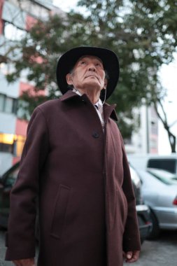 Şehir ortamında kulaklıkla müzik dinlerken şapka ve palto giyen yaşlı bir adam yukarı bakıyor.