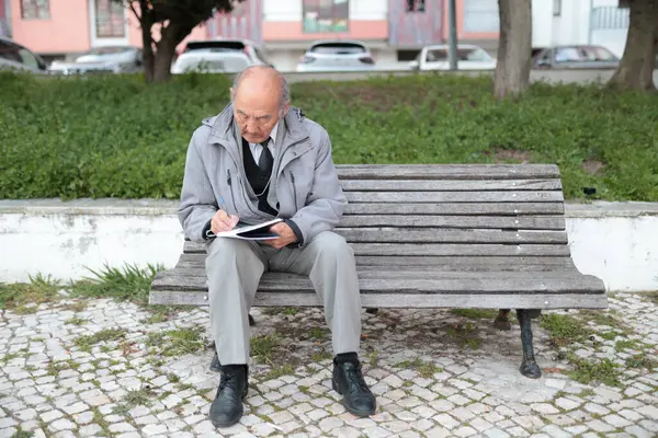 Yaşlı profesör park bankında otururken deftere not alıyor.