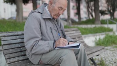 Kıdemli adam parkta boş zamanlarının tadını çıkarıyor, kulaklıkla müzik dinlerken bir deftere yazı yazıyor.
