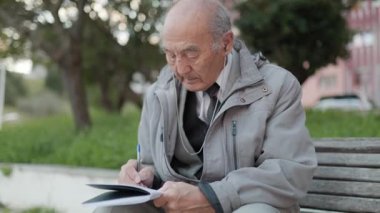 Kıdemli adam bir parkta bankta otururken defterine not alıyor.