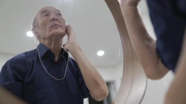 Kıdemli adam aynaya bakarken işitme cihazını ayarlıyor.