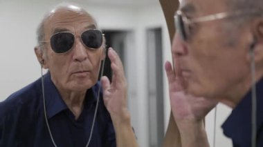 Yaşlı adam aynaya bakarken saçlarını ve güneş gözlüğünü düzeltiyor.