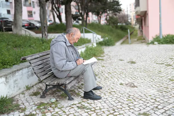Yaşlı bir adam parktaki bankta otururken bir kağıt parçasına notlar yazıyor.