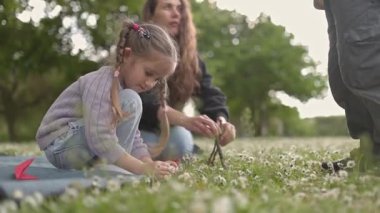 Anne ve çocuklar parkta papatya zinciri yapıyorlar.