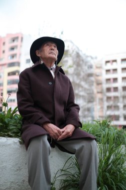 Şehir ortamında paltolu ve şapkalı son sınıf öğrencisi dışarıda oturuyor, yukarı bakıyor.