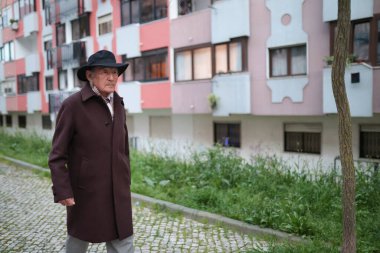 Apartman binasının yanındaki kaldırım taşı sokakta yürüyen şapka ve palto giyen son sınıf öğrencisi.