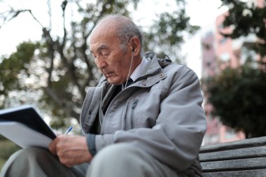 Yaşlı profesör, parkta bir bankta kulaklıkla müzik dinlerken not alıyor.