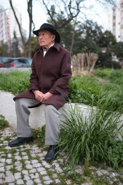 Üst düzey bir adam şapka ve ceket giyer ve dışarıda alçak duvarda oturup eğlenir.