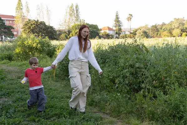 Anne ve oğlu uzun otların arasında rahatça geziniyor, el ele tutuşuyor ve iç açıcı bir an yaratıyorlar.