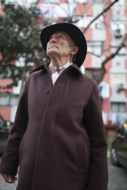 Şehir ortamında müzik dinlerken şapka ve palto giyen yaşlı bir adam yukarı bakıyor.