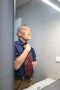 Zarif kıdemli işadamı, banyo aynasına yansıyarak, işitme cihazı takarken kravatını düzeltiyor.