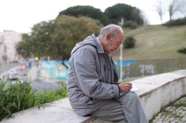 Yaşlı bir adam kulaklıklı müzikten hoşlanıyor ve şehir parkındaki bir deftere notlar yazıyor.